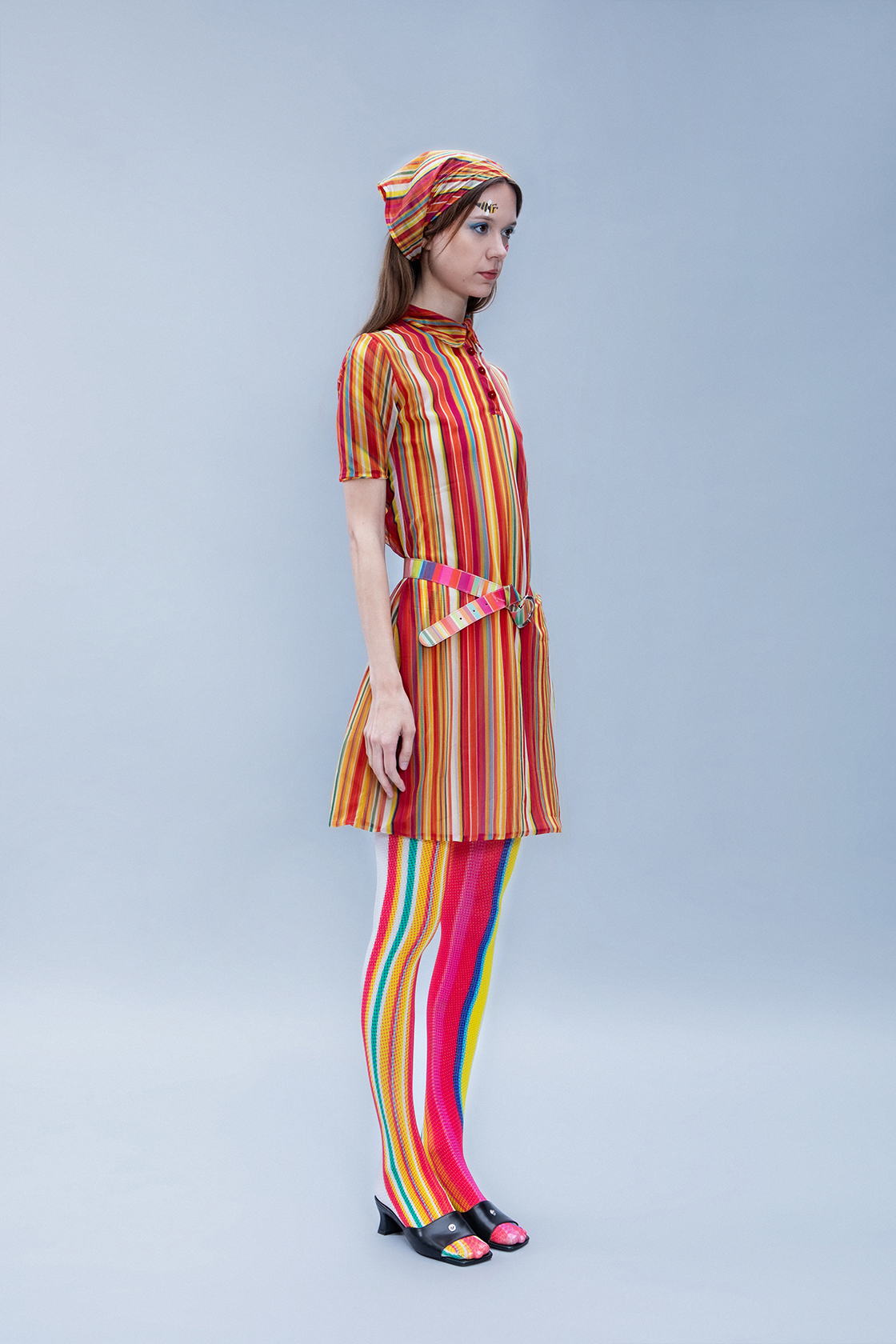 Full body shot of a model in colorful striped dress, scarf and leggings facing 3/4 away from the camera against a gray backdrop